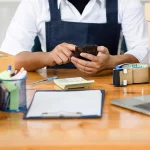 A small business owner managing online orders using a smartphone and laptop, surrounded by packaging materials.