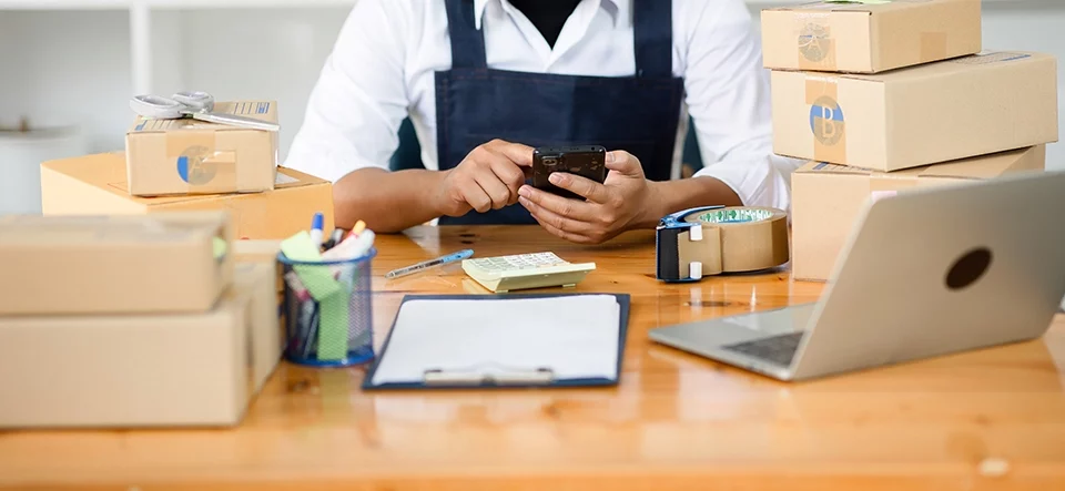 A small business owner managing online orders using a smartphone and laptop, surrounded by packaging materials.